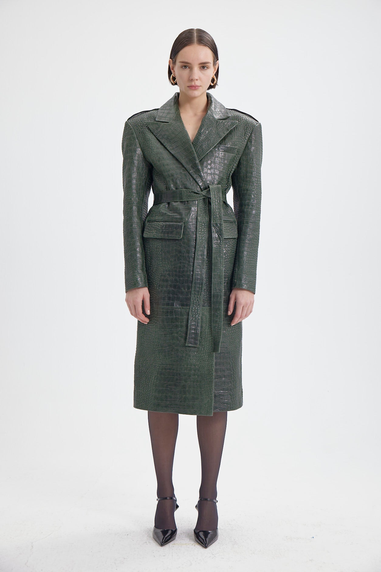 Elegant green croco-pattern leather coat with a belt and front flap pockets, modeled by a woman.