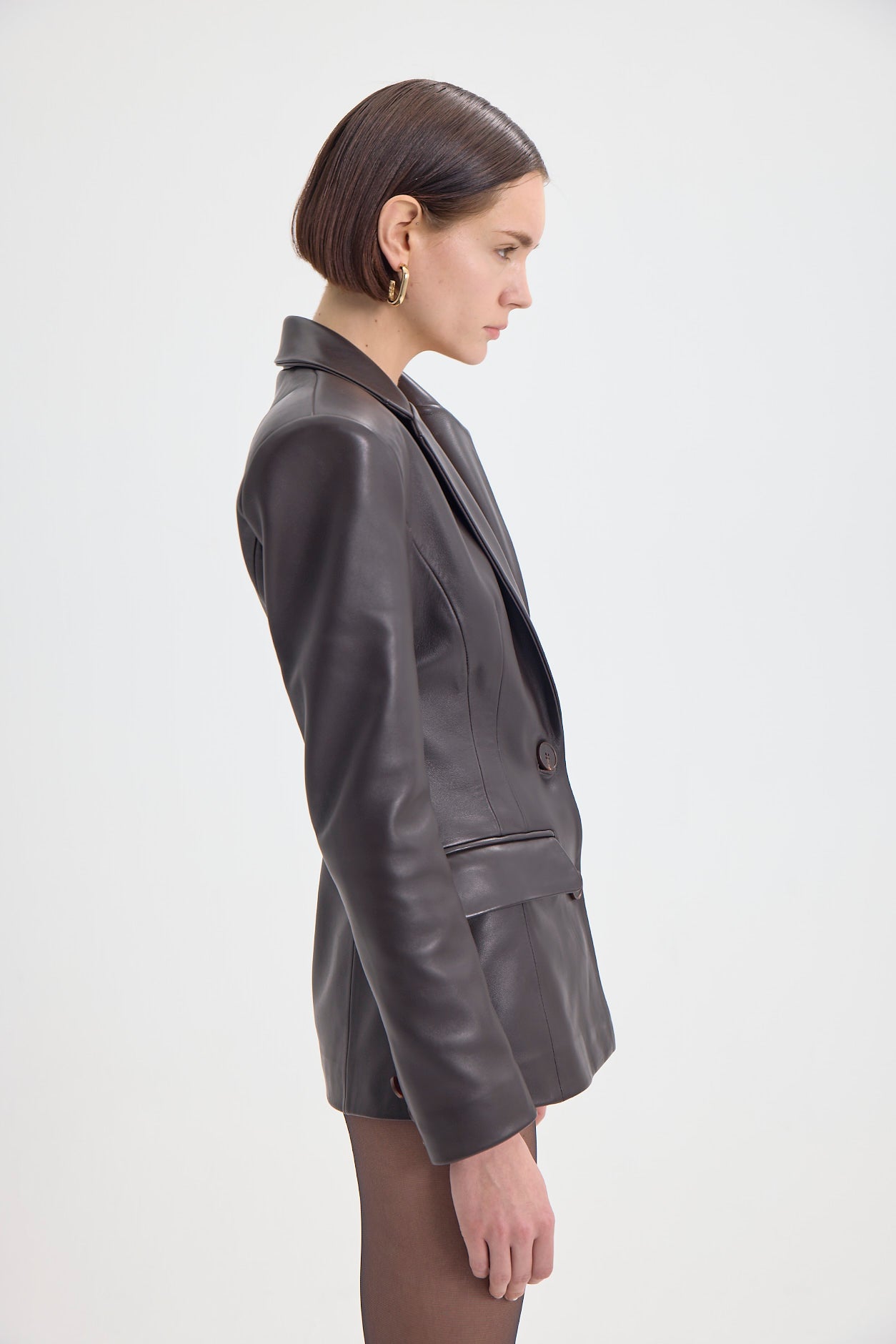 Woman in a sleek dark brown leather blazer featuring a notched lapel and front flap pockets, side profile.