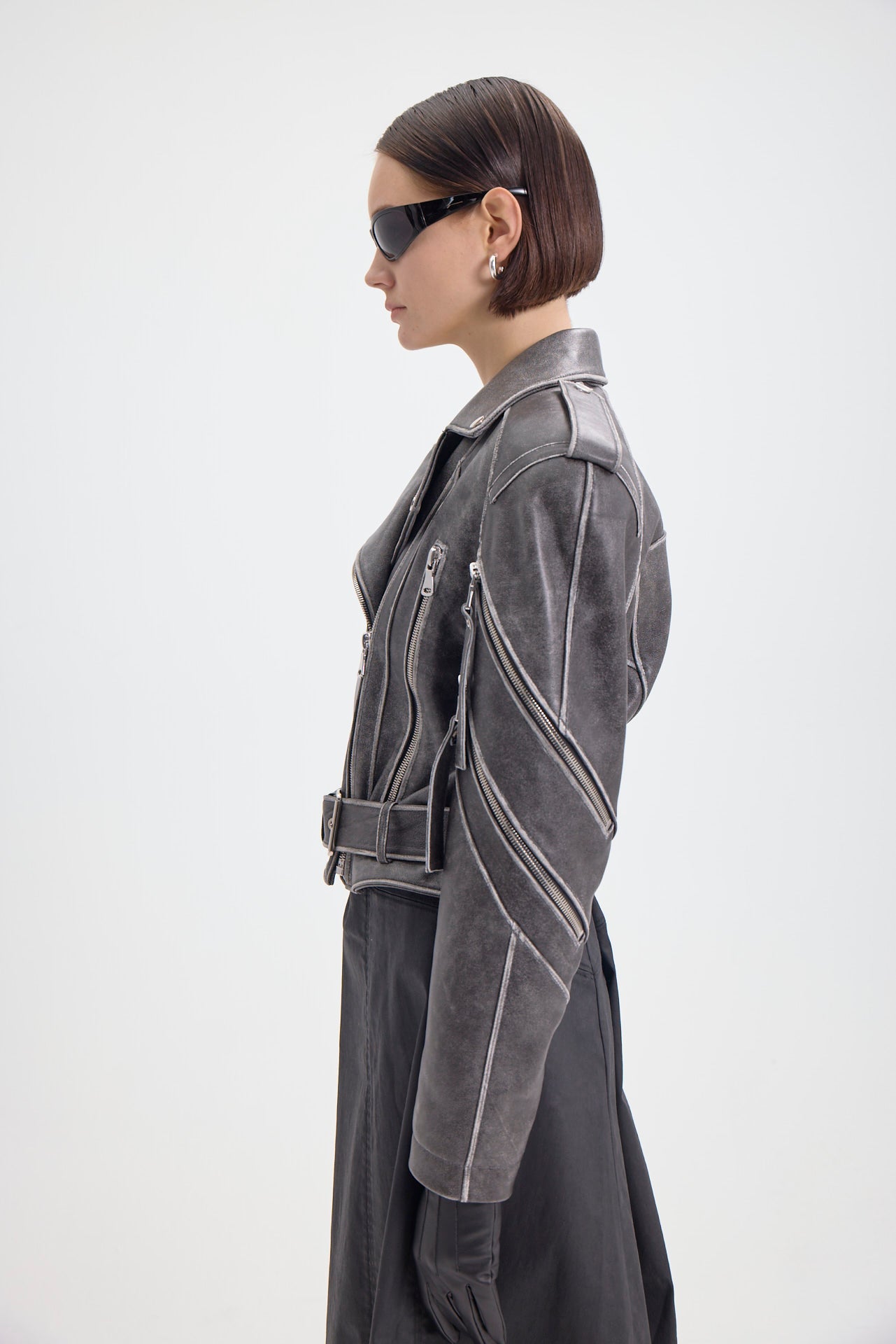 Woman in a gray leather biker jacket with multiple zippers and a belted waist, viewed from the side.